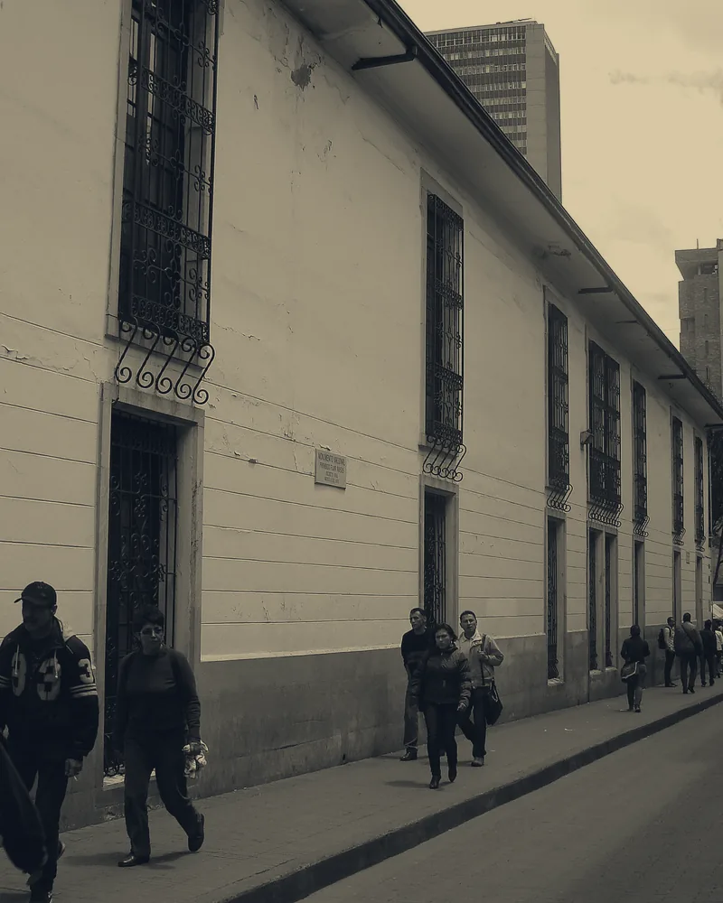 Calle del centro histórico de Bogotá junto a un muro blanco con ventanas de reja, con personas caminando